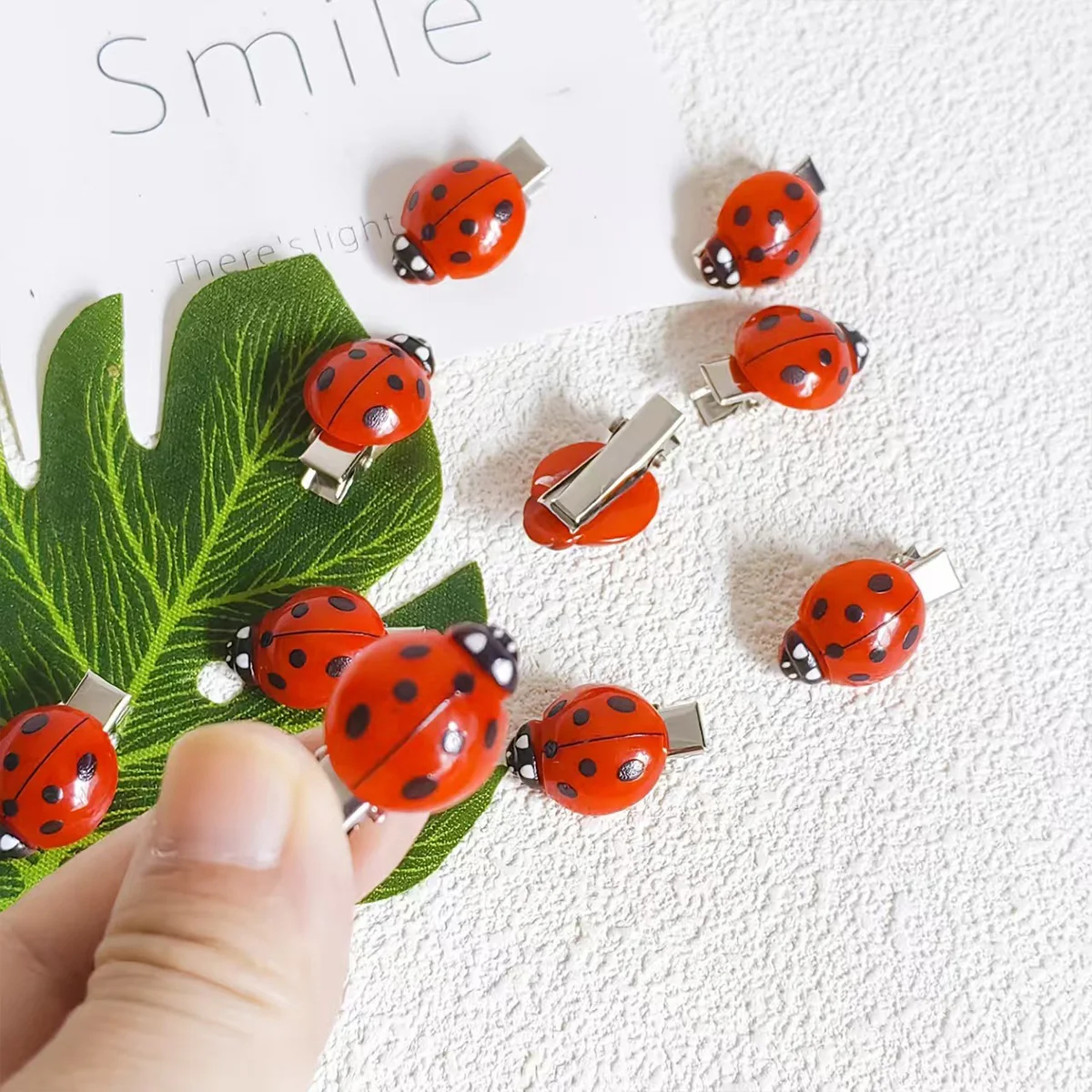 Red ladybug-shaped hair clips on a textured white background with a green leaf.