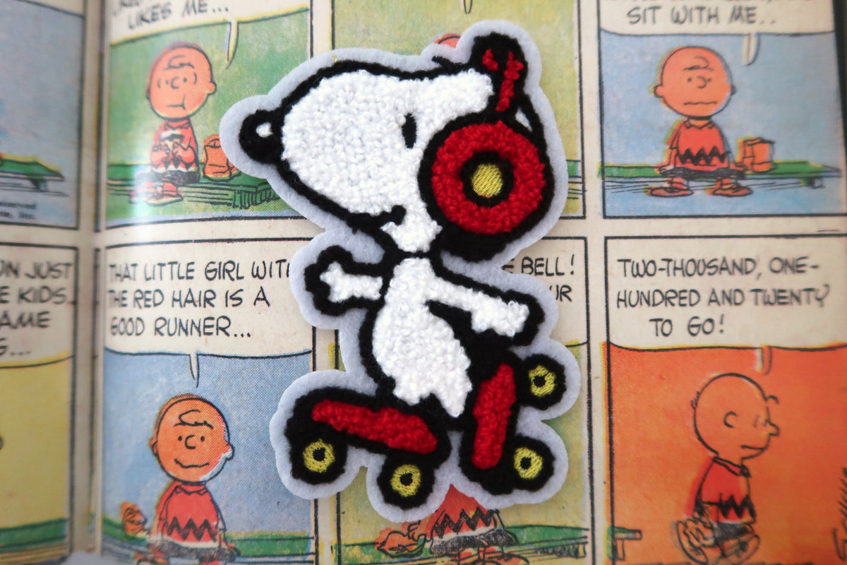 A Snoopy embroidered patch on a red skateboard with headphones, displayed on a background of a Peanuts comic book page.