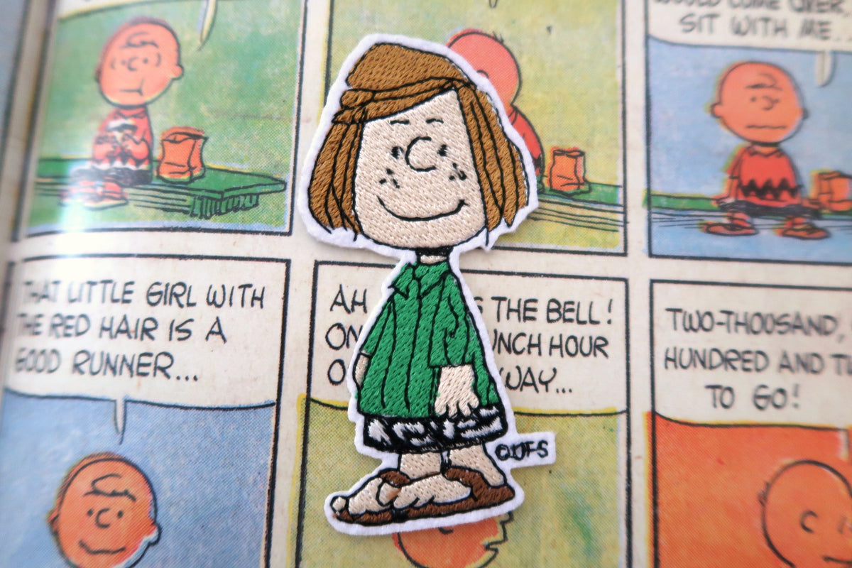 A die-cut sticker of the character Peppermint Patty from Peanuts, featuring her green shirt, brown hair, and black-and-white skirt, placed on top of a printed Peanuts comic page.