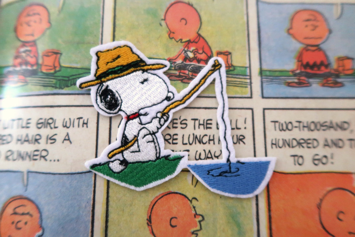 A close-up of a embroidered patch featuring Snoopy fishing on a comic-style background with Peanuts characters and panels.