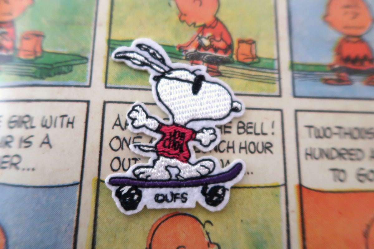 A close-up of a Peanuts Snoopy embroidered patch riding a purple skateboard, placed on top of a vintage-style comic book page featuring Charlie Brown.
