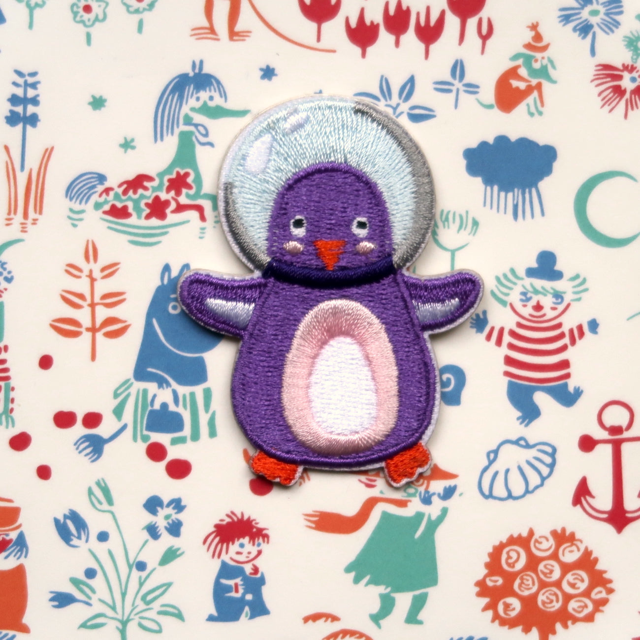 A purple and orange embroidered sticker patch of an astronaut penguin on a background with various colorful patterns.