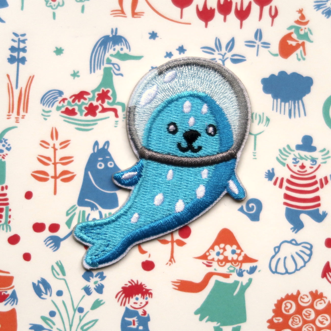 An embroidered sticker patch of an astronaut seal in blue and white colors with a background pattern of various playful illustrations including animals and astronauts.