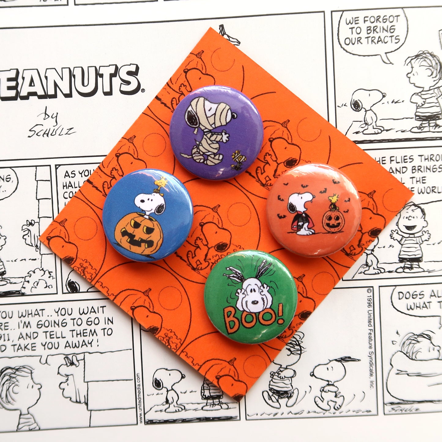 A set of four vintage style Halloween themed pin badges featuring characters from the Peanuts comics, placed on an orange background with a comic strip pattern.