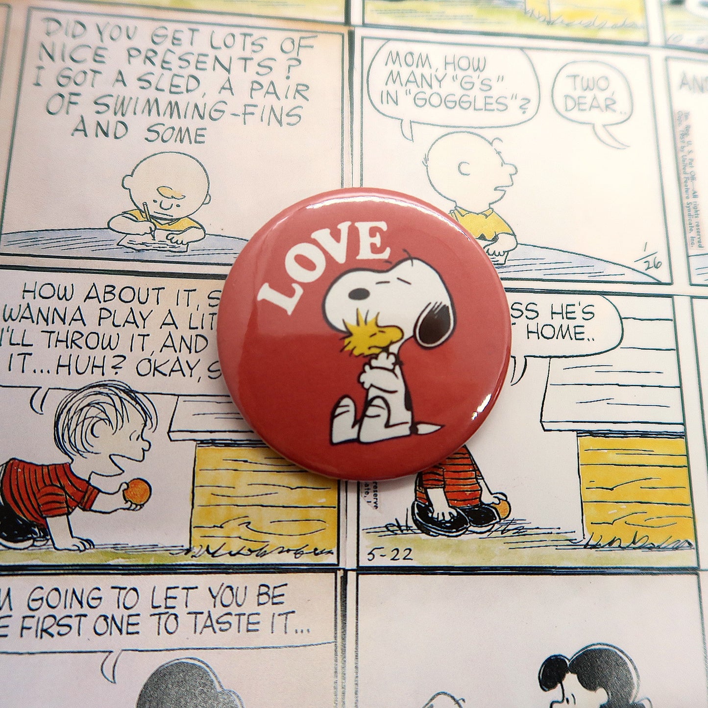 A vintage can badge featuring Snoopy and Woodstock with the word 'LOVE' on a red background, displayed over a comic strip.