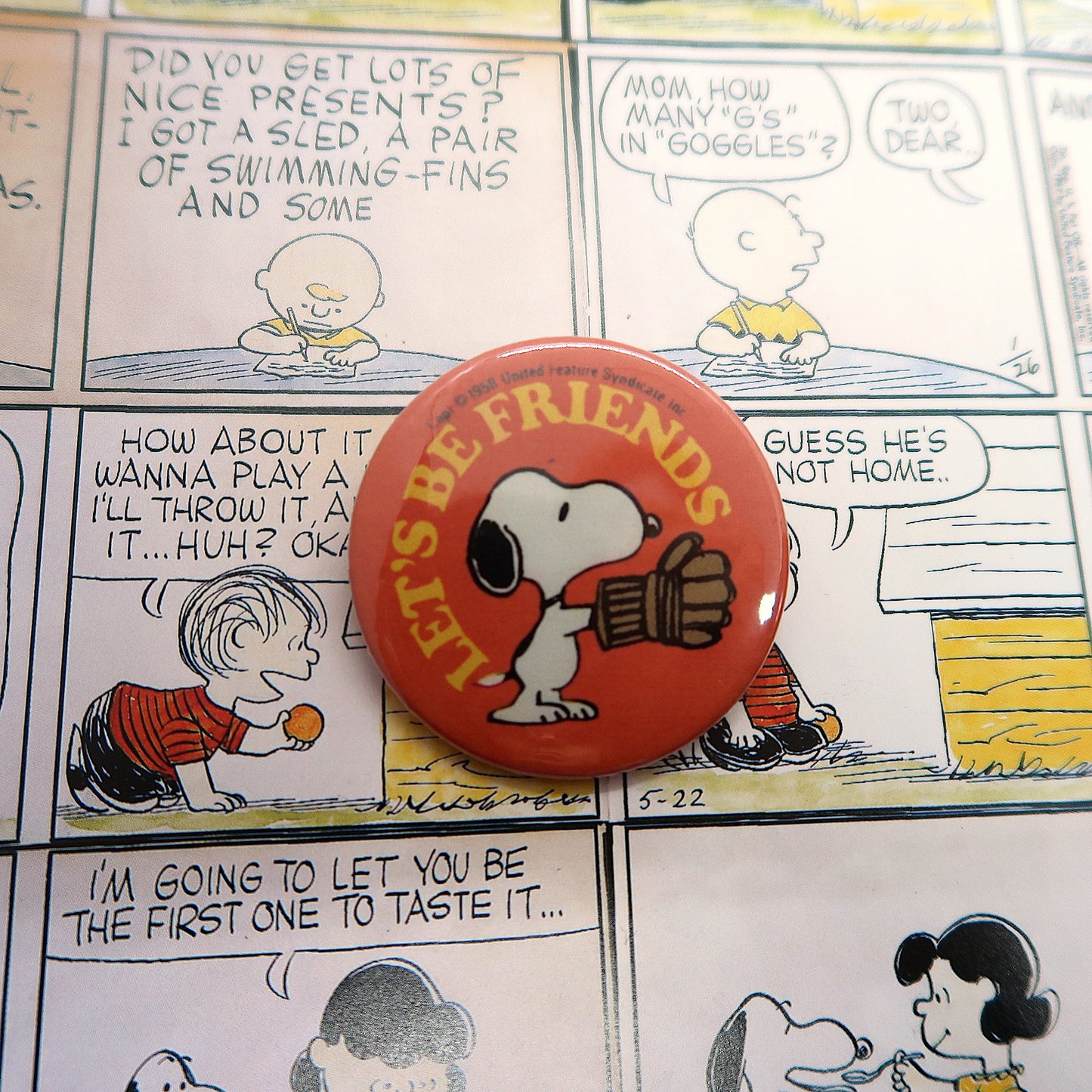 A vintage-style pin badge featuring the character Snoopy from the Peanuts comics, with the text 'Let's Be Friends' on it, displayed over a comic strip background.
