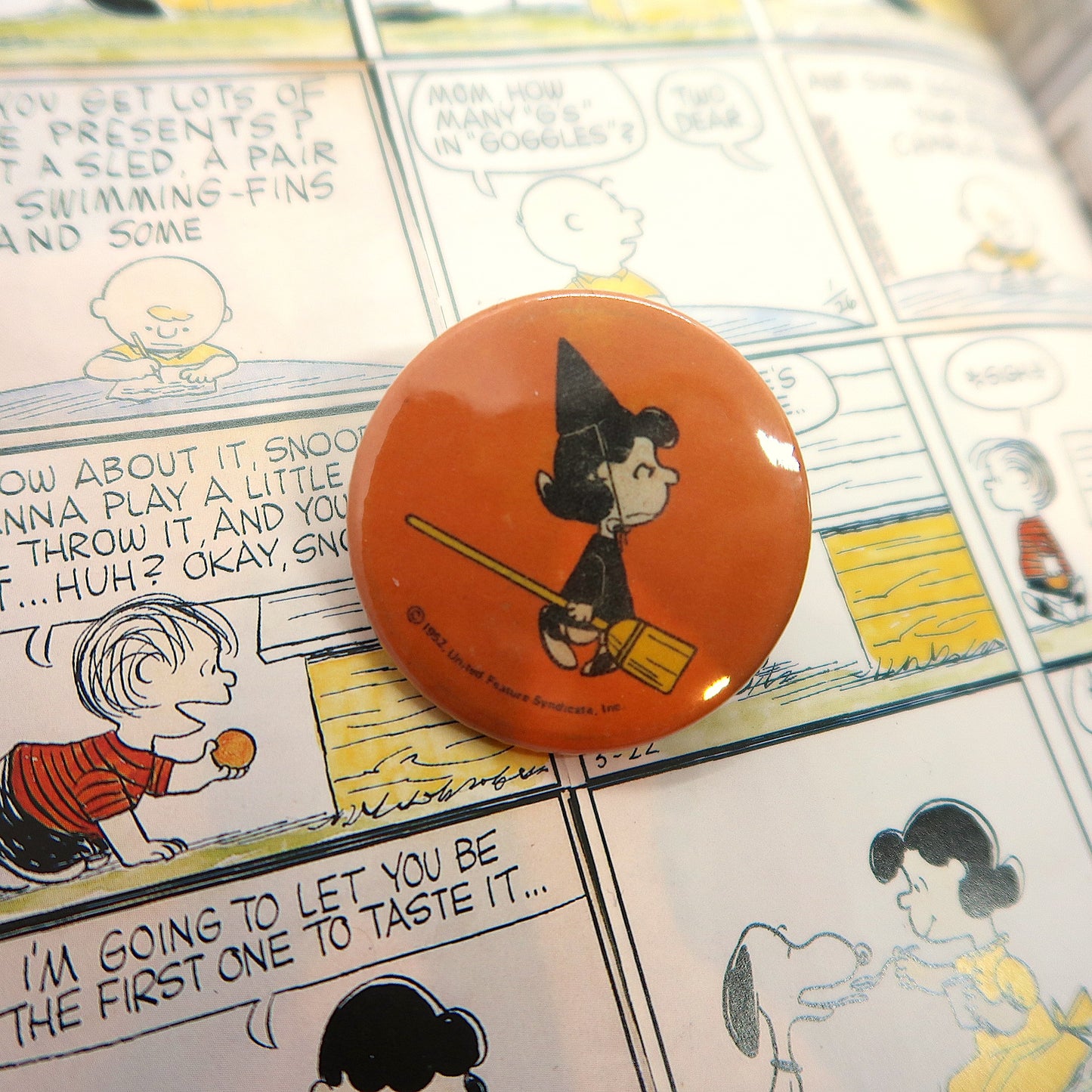 A vintage badge featuring Lucy van Pelt from the Peanuts comics dressed as a witch, with an orange and black color scheme, found on a background depicting a comic strip.