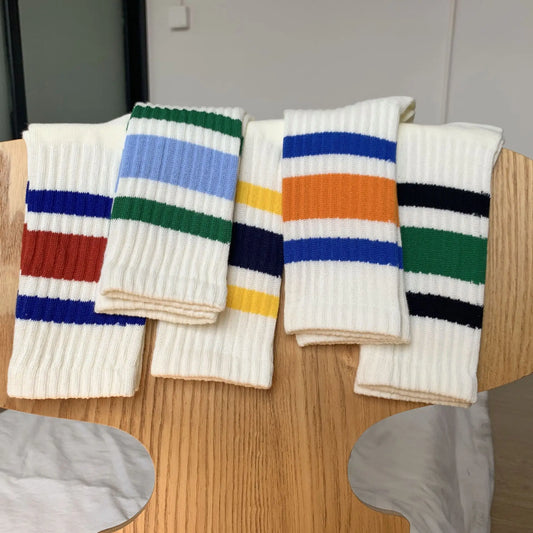 Five pairs of white ribbed knit socks with colorful horizontal stripes are arranged on a wooden table. The socks feature various stripe combinations including blue, green, orange, red, yellow, and black.