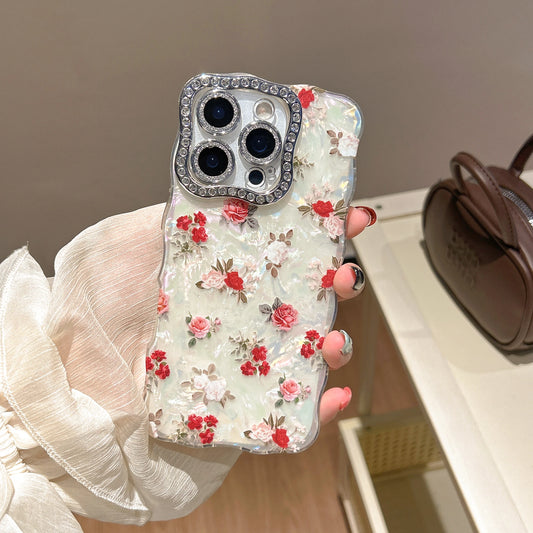 A hand wearing a white sheer sleeve holds a clear phone case decorated with red and pink 3D roses on a light green background, featuring a rhinestone-embellished camera frame.