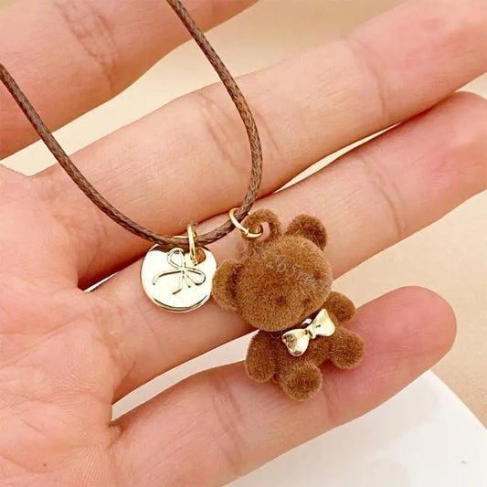 A close-up of a hand holding a delicate brown cord necklace with a small brown teddy bear charm and a gold-toned round pendant.