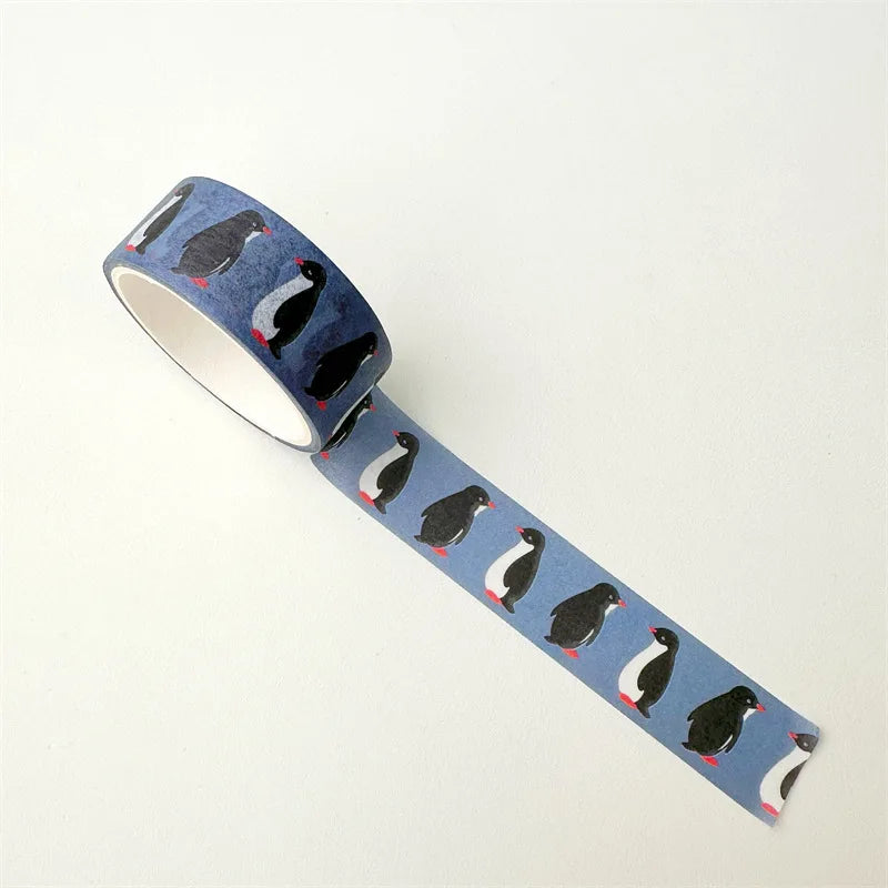 A roll of washi tape featuring a repeating pattern of penguins on a light blue background, with black, white, and red accents, shown on a plain off-white background.