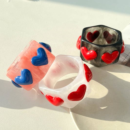 Three translucent resin bangles with raised heart details, displayed on a light surface in bright natural light. One ring is peach-pink with blue hearts, one is white with red hearts, and one is smoky gray with red hearts.