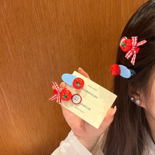 A person holds a pair of light blue hair clips decorated with red tomatoes and red-and-white gingham bows, while wearing matching clips in their hair. The background is a warm wood panel wall.