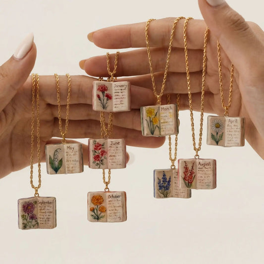 Hand holding necklaces with small book pendants featuring floral designs and months of the year text.