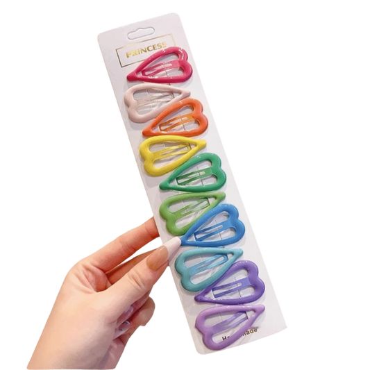 A hand holds a white card displaying ten pastel-colored heart-shaped hair clips in a vertical row. The clips come in pink, light pink, orange, yellow, green, blue, teal, purple, and navy.