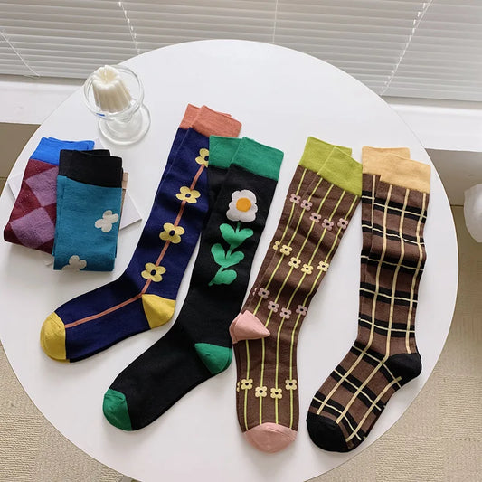 Four pairs of patterned knee-high socks arranged on a round white table. The socks feature colorful floral, geometric, and plaid designs in navy, black, brown, blue, teal, green, pink, yellow, and orange.