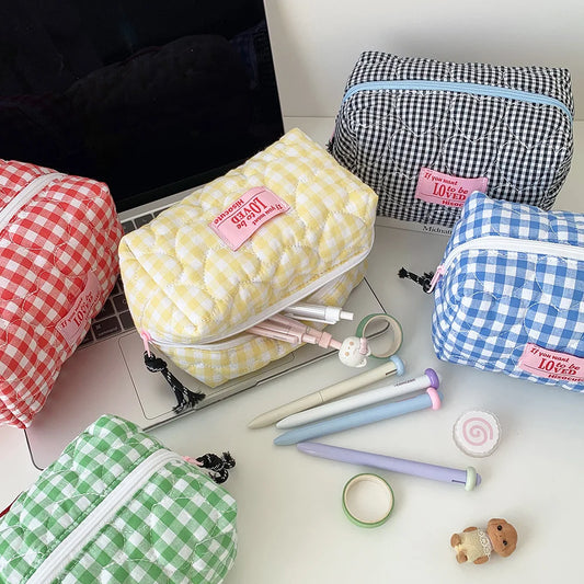 Five quilted zippered pencil cases in different gingham patterns (red, yellow, blue, green, and black-and-white) arranged on a desk with stationery items.