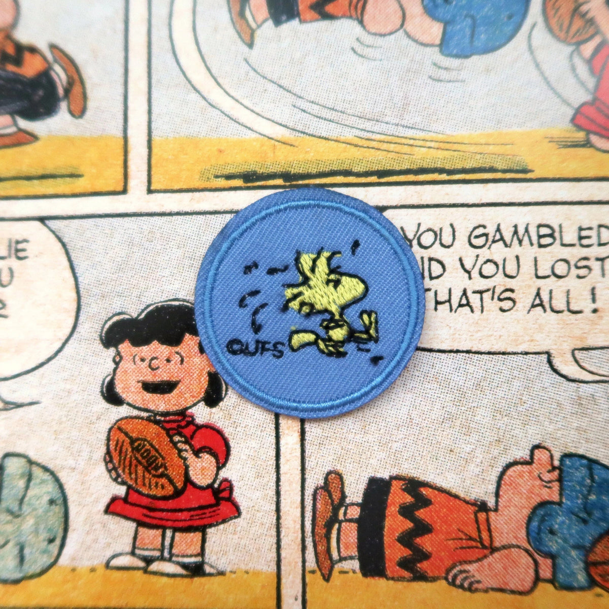 A circular embroidered patch featuring Snoopy and Woodstock on a blue background, placed on a printed Peanuts comic panel with cartoon characters and speech bubbles.