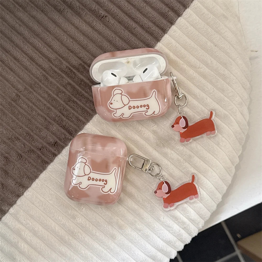 A pair of translucent pink AirPods cases with cartoon dachshund illustrations, shown open and closed with matching acrylic keychain charms on a ribbed fabric surface.