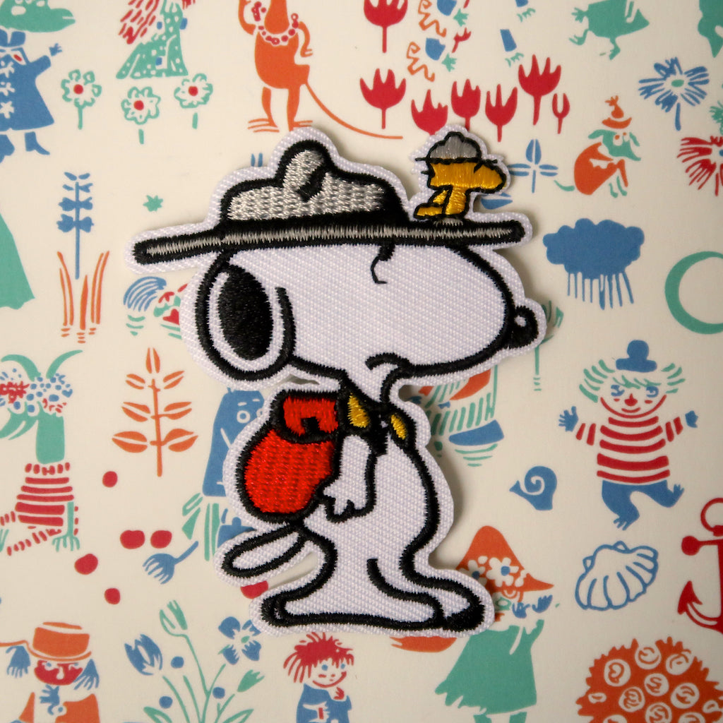 An embroidered patch featuring Snoopy from the Peanuts comic strip with a boyscout uniform, a hat with Woodstock the bird, and a backpack.
