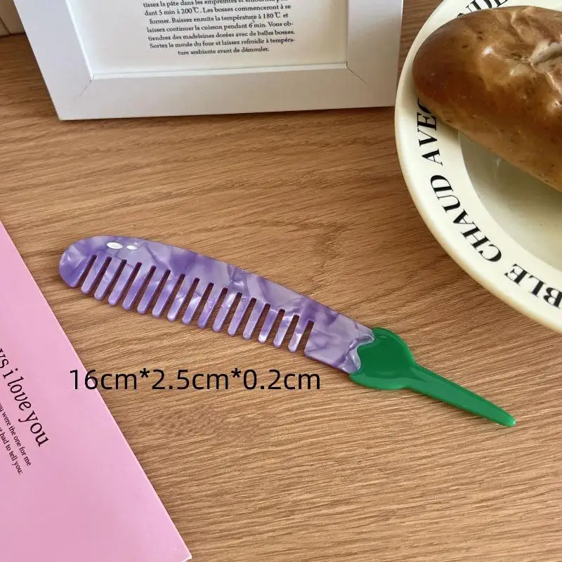 Purple eggplant comb with green handle on a wooden surface next to a pink book and a bagel.