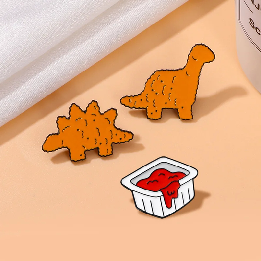 A set of three orange enamel pins with dinosaur nugget designs and a small container of ketchup depicted alongside.