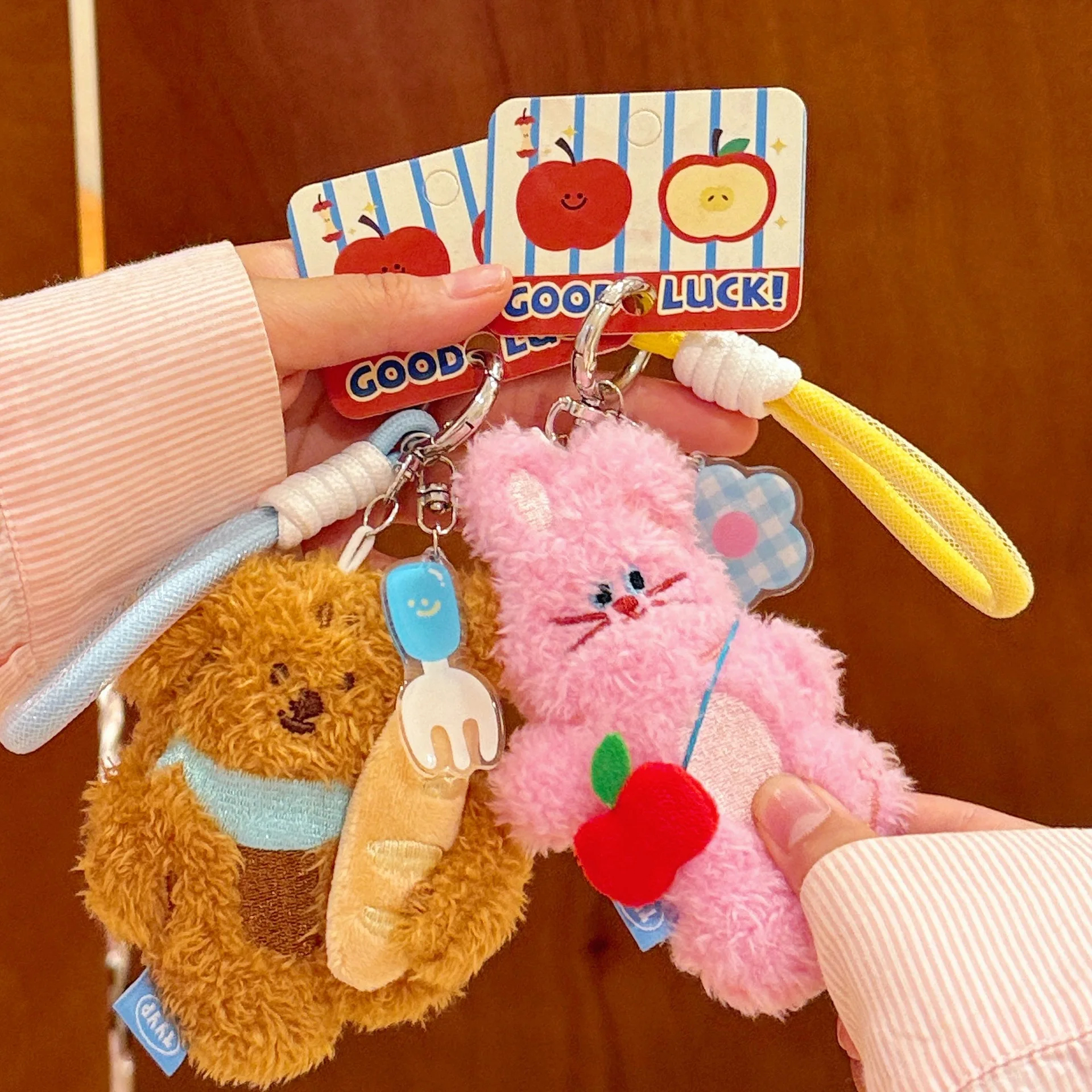 Two plush keychains, one brown bear and one pink bunny, held against a wooden background with a 'Good Luck!' card.