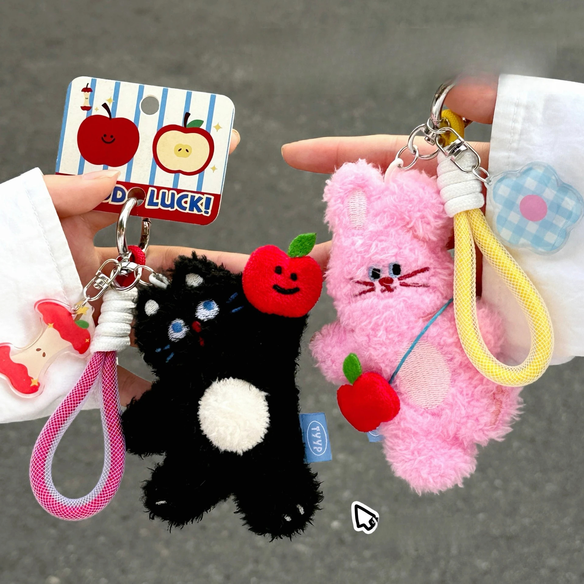 Two plush keychains, one black and white, one pink, held by a hand with a gray background.