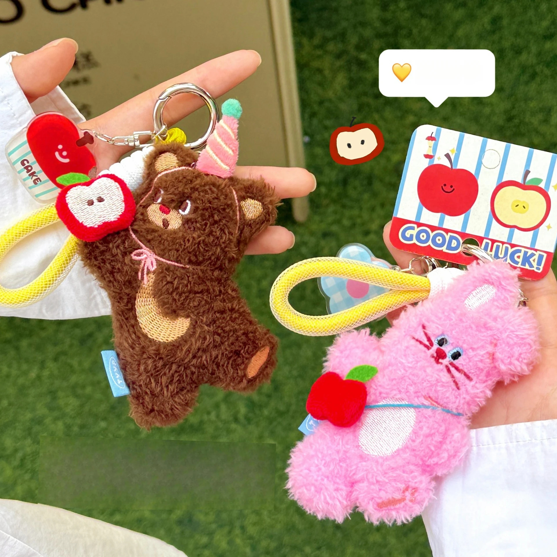 Two plush keychains, one brown bear and one pink cat, held by a person with a green background.