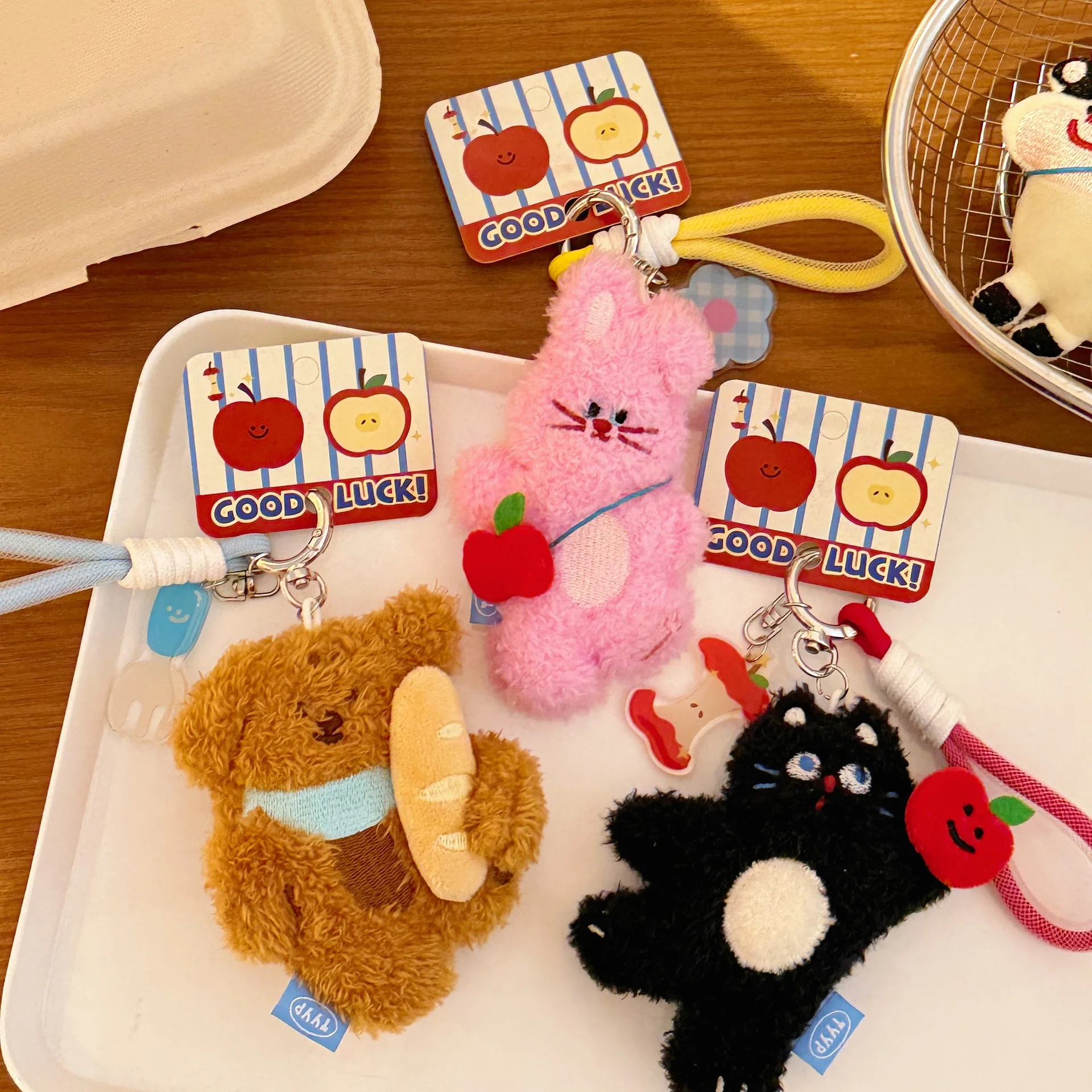 Plush animal keychains shaped like a black and white cat, a pink bunny and brown puppy with a baguette with 'Good Luck!' cards on a wooden surface