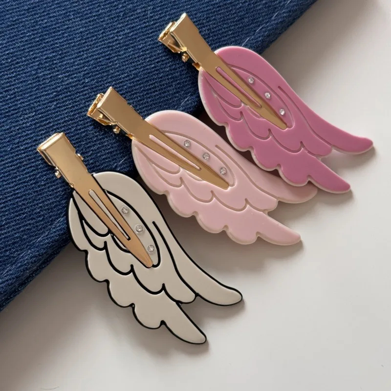 Three angel wing hair clips in pink, white, and gold on a blue and white background.