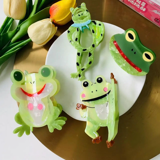 A set of four acrylic hair claw clips in the shapes of green frogs in different poses, displayed on a golden plate.