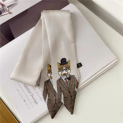 A cream-colored silk necktie with a printed graphic of two cat characters dressed in brown plaid suits and top hats, laid flat on top of an open book.
