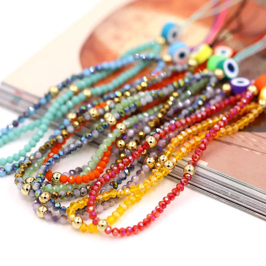 A close-up of multiple strands of colorful beaded wrist straps arranged on a light surface, featuring small round beads in turquoise, blue, green, orange, red, yellow, purple, and metallic gold, with evil eye beads near the top.