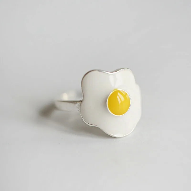 A novelty ring designed to look like a fried egg, featuring a white enamel-filled flower-like shape with a bright yellow yolk in the center and a silver-tone metal band, set against a plain light gray background.