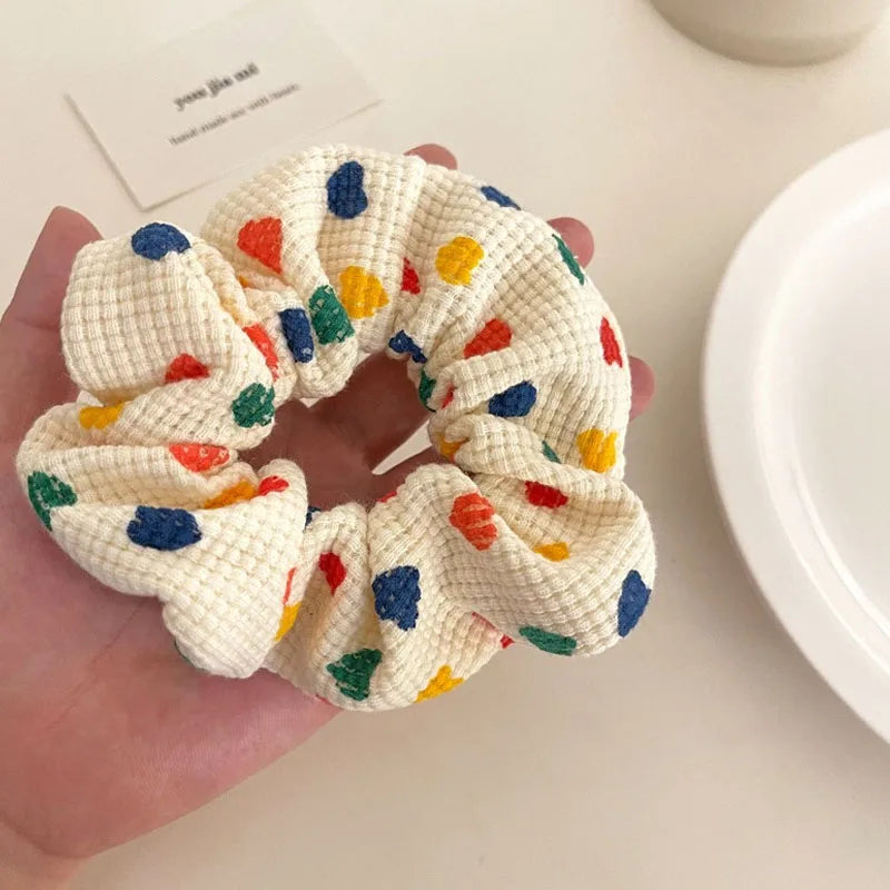 A hand holds a cream-colored textured fabric scrunchie decorated with colorful embroidered polka dots in blue, green, yellow, and orange, set against a softly blurred light beige background with a white plate.