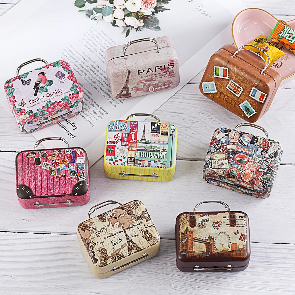 Seven small square cosmetic cases with vintage suitcase and Paris-themed prints are arranged on a white wood surface with an open book and flowers in the background. The cases feature different colorful patterns including pink floral, beige Eiffel Tower, yellow Paris collage, pink striped, multicolor postage stamp, cream map, and dark brown cityscape designs.