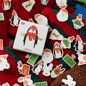 A white square box labeled 'Life to Sticker' with a penguin illustration, surrounded by many small Christmas-themed sticker cutouts including Santa, penguins, gifts, and snowman shapes on a red textured fabric background.