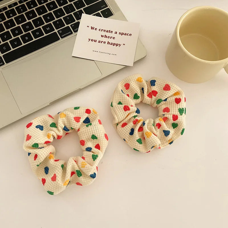 Two cream-colored textured fabric scrunchies with colorful heart patterns are displayed on a white desk alongside a laptop, a card with an inspirational quote, and a beige ceramic mug.