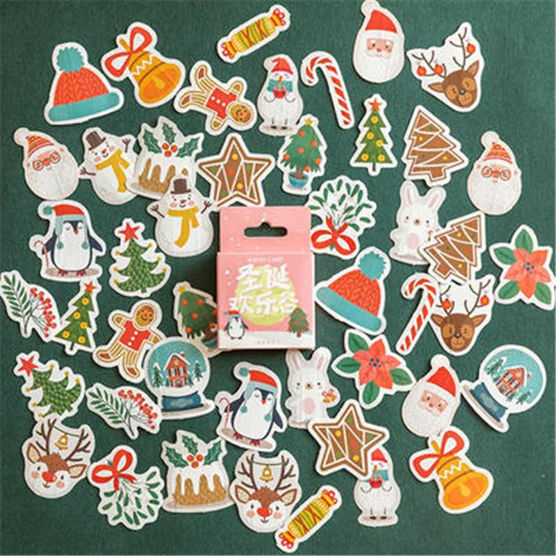 A set of colorful Christmas-themed sticker cutouts arranged around a small pink hanging card on a dark green background. The stickers include Santa Claus, penguins, Christmas trees, candy canes, reindeer, snow globes, bows, bells, stars, gingerbread houses, and holiday foliage.