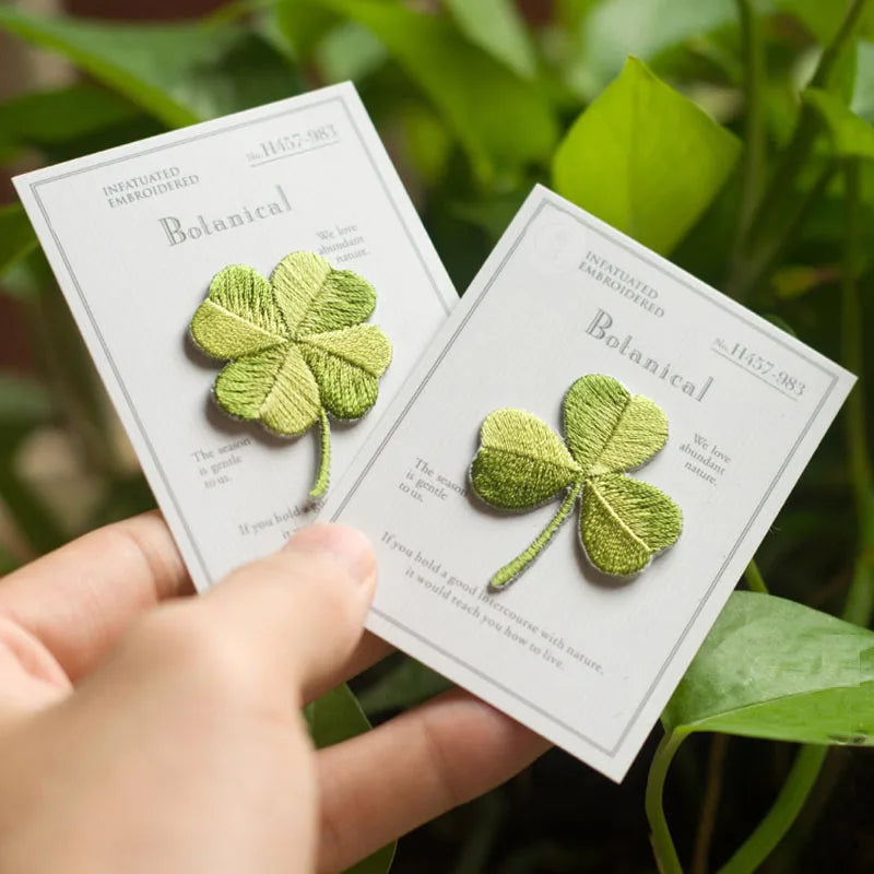 A hand holds two small embroidered clover patches on printed cards in front of green leaves. The cards read 'Botanical' and 'Unfinished Embroidered,' and the patches are light green with stitched leaf details.