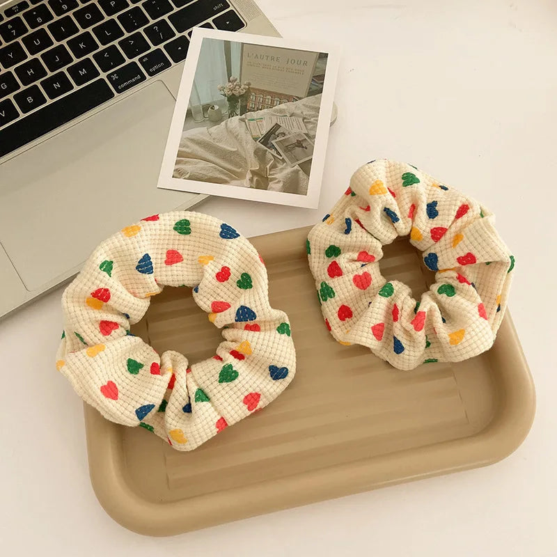 Two cream-colored waffle-knit fabric scrunchies with a colorful heart pattern, displayed on a beige tray next to a laptop and a photo print.