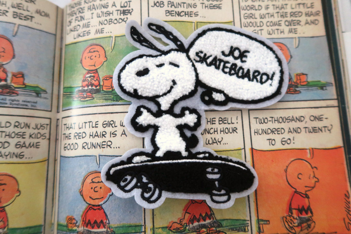 A Snoopy embroidered patch on a skateboard, featuring a white and black design with a speech bubble that reads 'JOE SKATEBOARD!'. The patch is displayed flat on top of a vintage Peanuts comic book page.