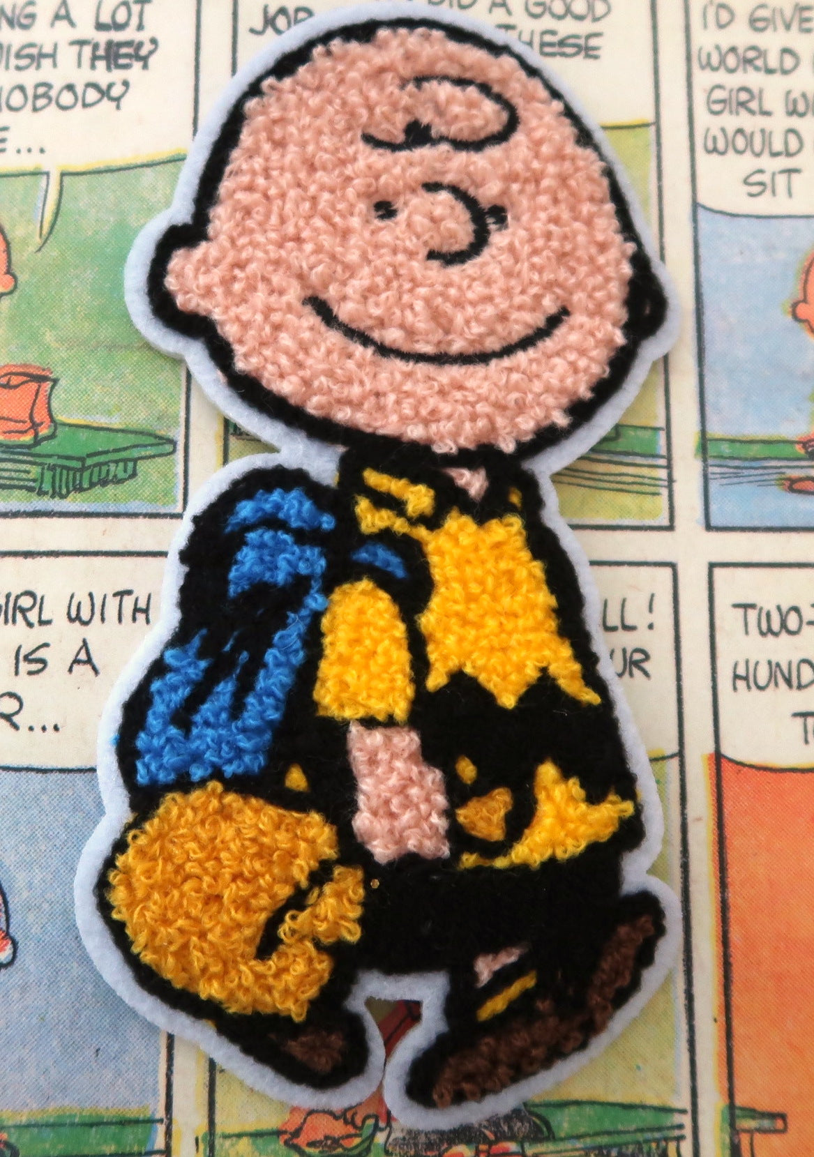 A close-up of a textured, looped fabric patch depicting the cartoon character Charlie Brown from Peanuts, shown against a background of vintage comic book pages.