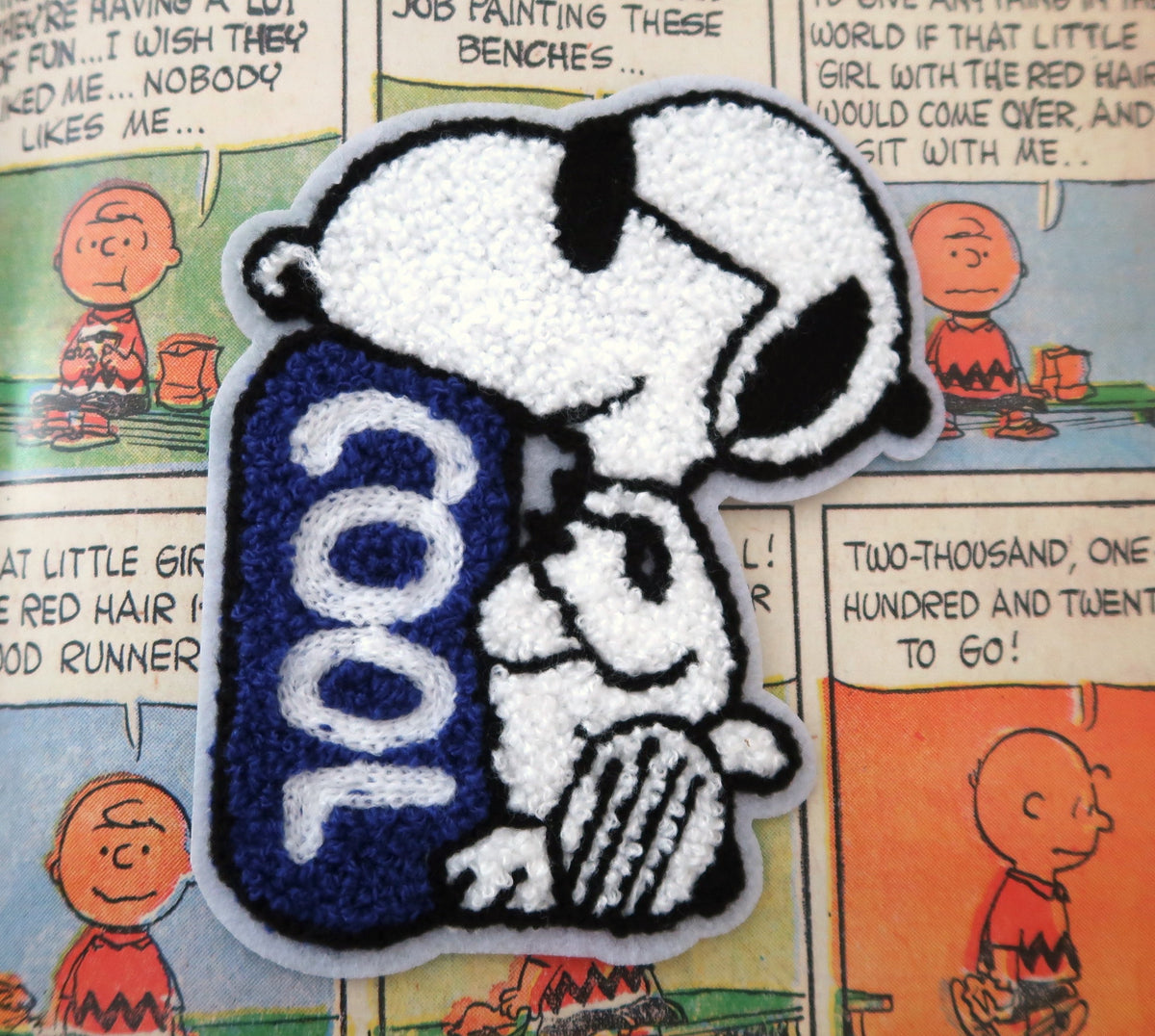 A Snoopy embroidered patch featuring Snoopy and a blue 'COOL' sign, placed on top of a printed Peanuts comic page.