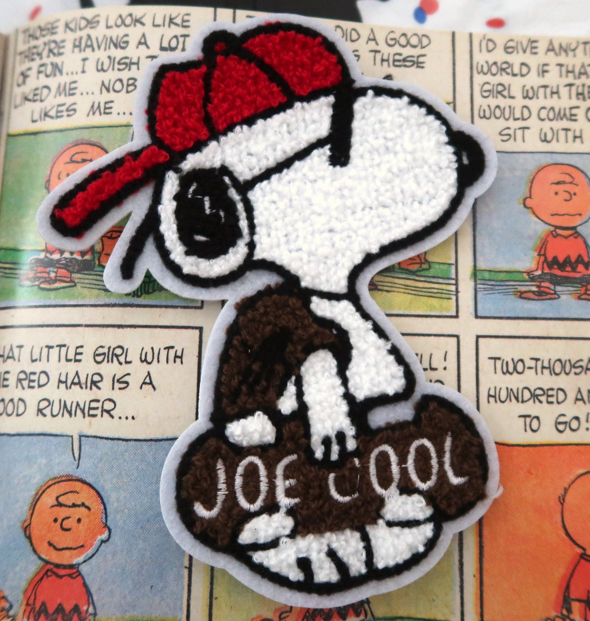 A close-up of a Snoopy embroidered patch wearing a red cap and holding a brown bag labeled 'JOE COOL', placed on top of a printed comic book page.