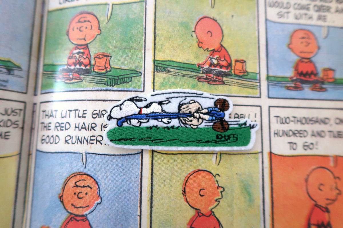 A die-cut patch featuring a Peanuts comic scene with Linus and Snoopy lying on a grassy field, placed on a printed Peanuts comic page.
