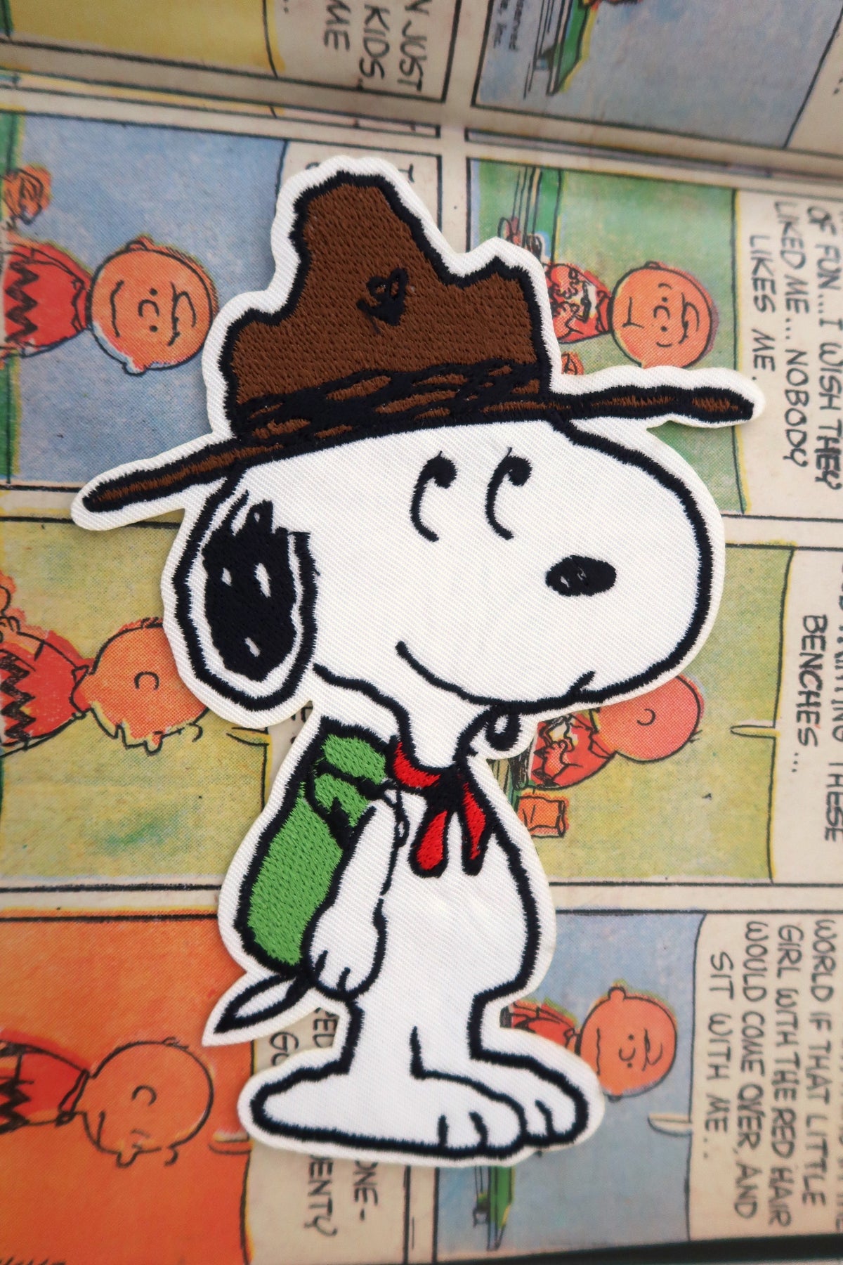 A die-cut embroidered patch of Snoopy wearing a brown scout hat, green backpack, and red neckerchief, placed on top of a Peanuts comic book page.