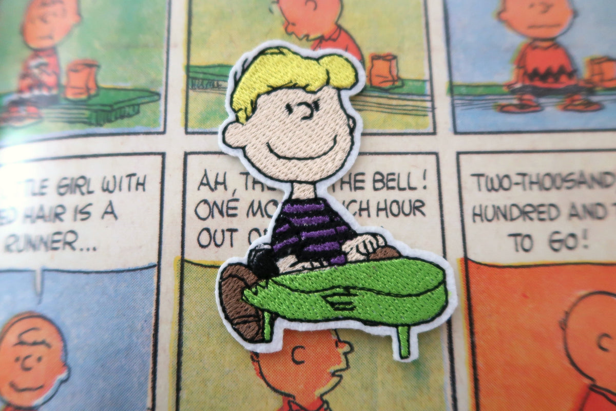 A close-up of a Peanuts character patch featuring Schroeder with a green toy piano, placed on top of a printed Peanuts comic page.