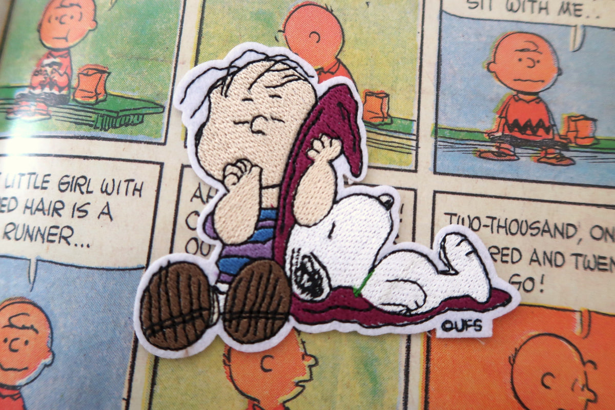 An embroidered patch featuring Snoopy and Linus from Peanuts, with a white border, placed on top of a printed Peanuts comic page.